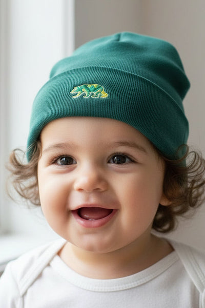 Child wearing a green dapper bear beanie with a logo, standing indoors near a window.